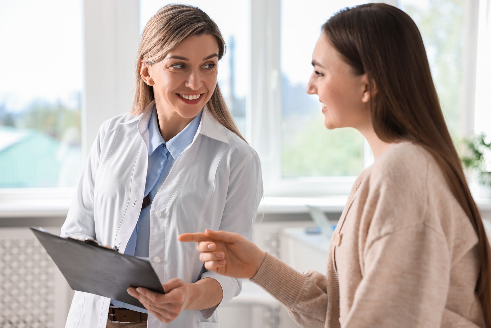 A medical professional in a white coat holds a clipboard and smiles while talking to a patient in an office setting.