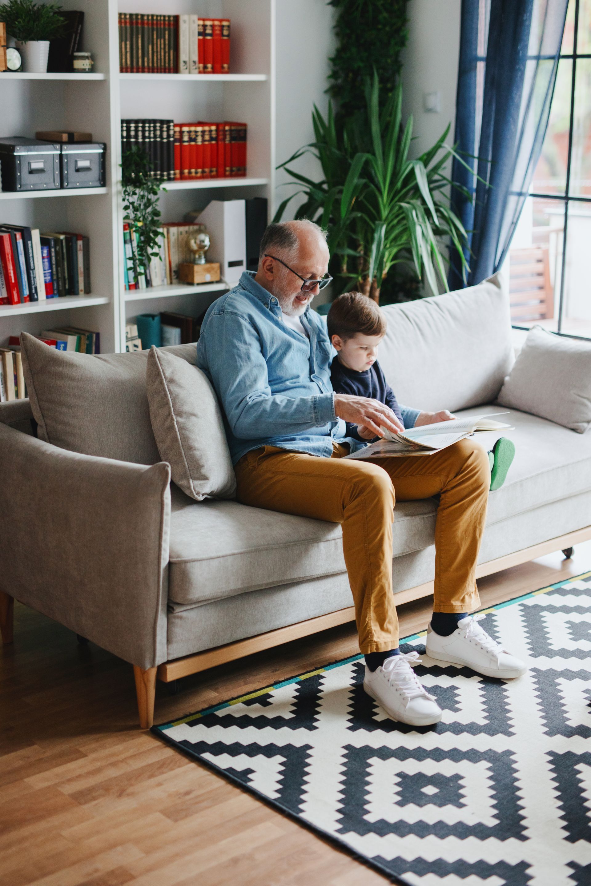 Grandfather and grandson reading a book together on a sofa in a living room.