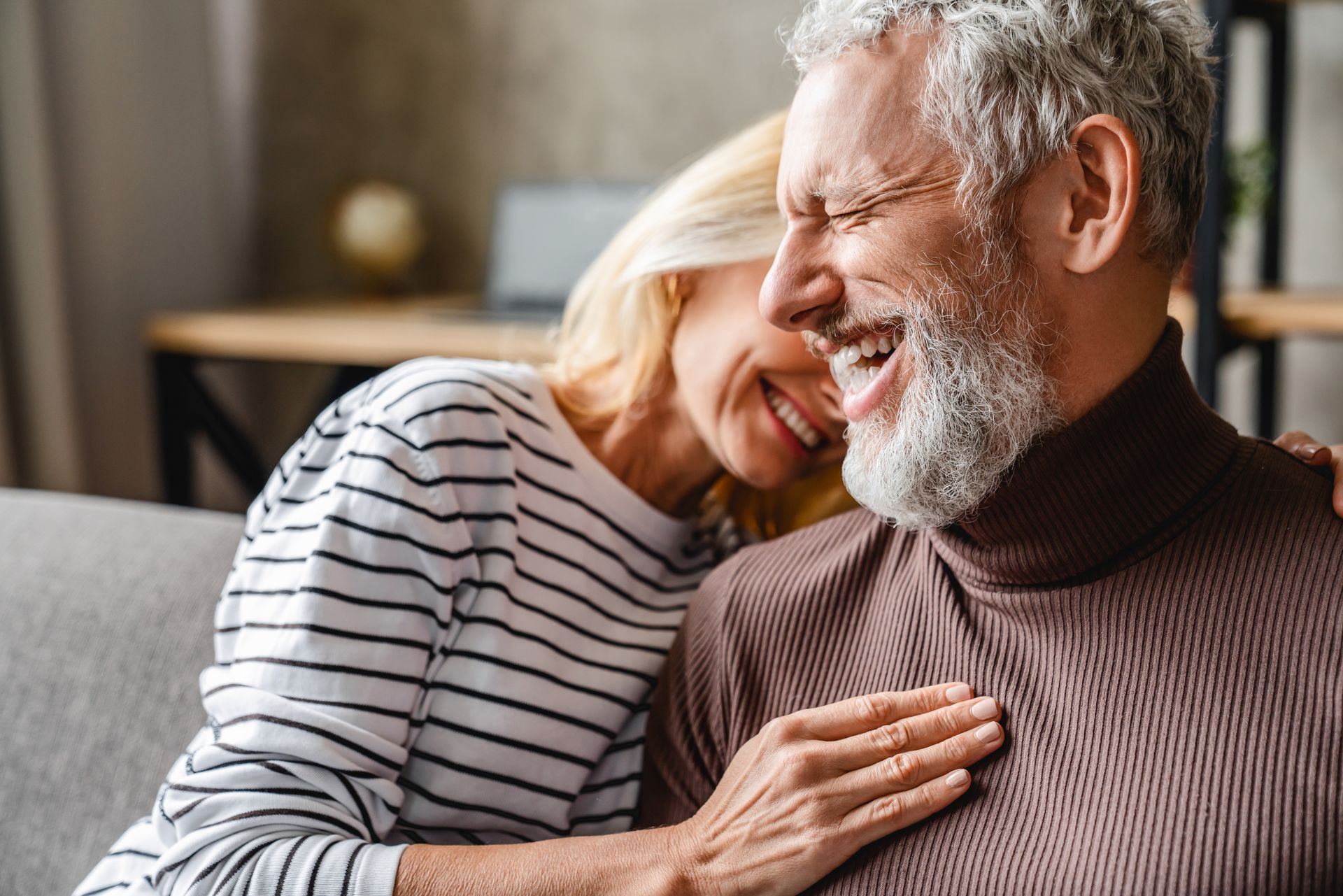Older couple laughing together, woman with blonde hair, man with gray beard, in living room.