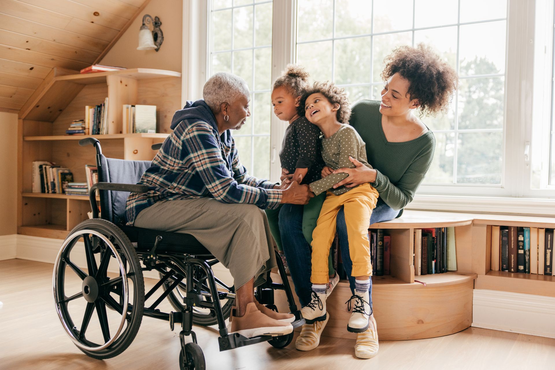 Family gathers around elderly person in wheelchair, near sunny window.