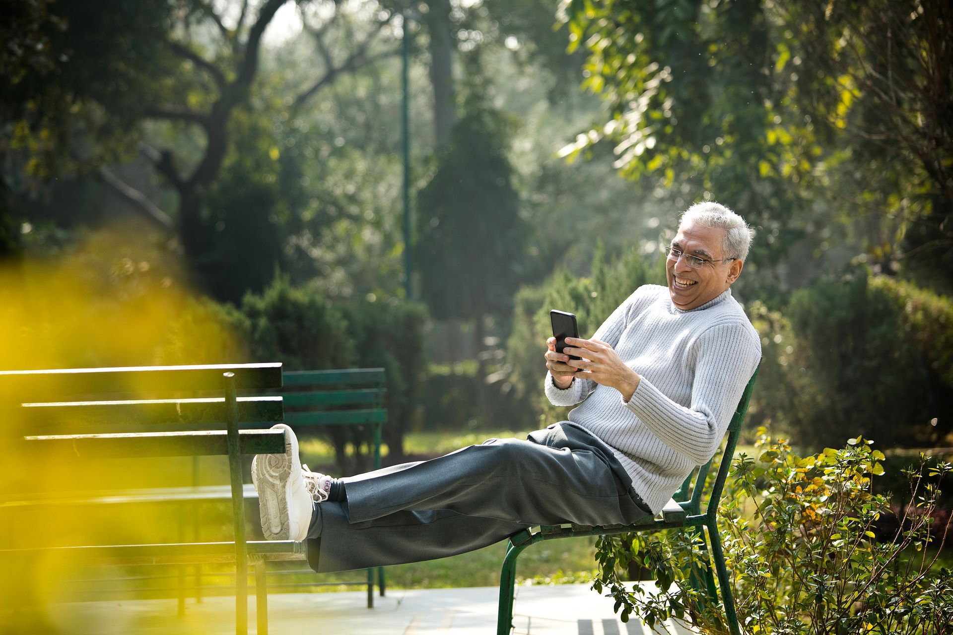 Smiling older man in glasses uses a phone while relaxing on a park bench outdoors.