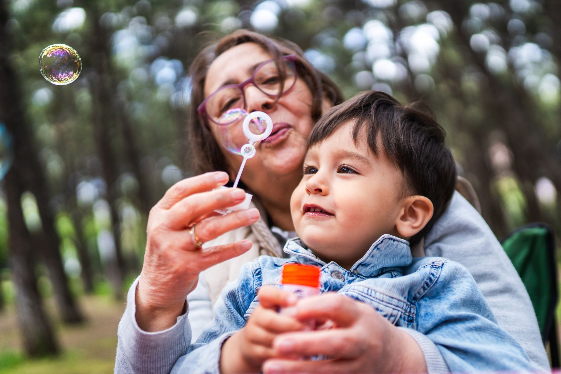Woman blowing bubbles for a smiling toddler in a park setting.