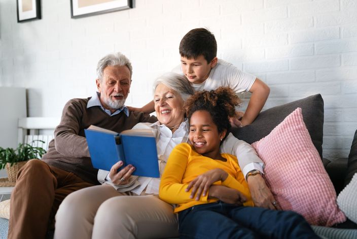 Grandparents and grandchildren read a book together on a sofa; everyone smiles.