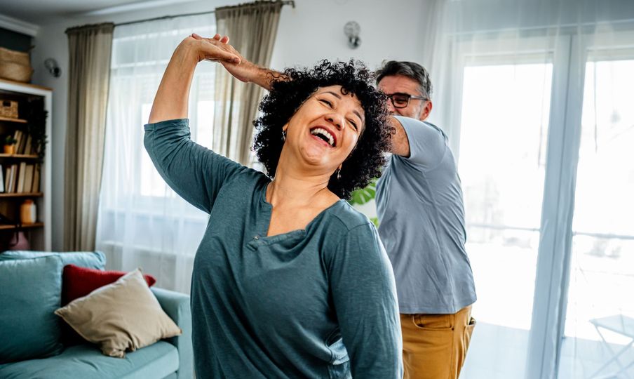 Couple dancing in a living room, woman laughing with arm raised, man behind her.