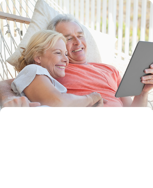 Senior couple smiles while relaxing in a hammock, looking at a tablet outdoors.