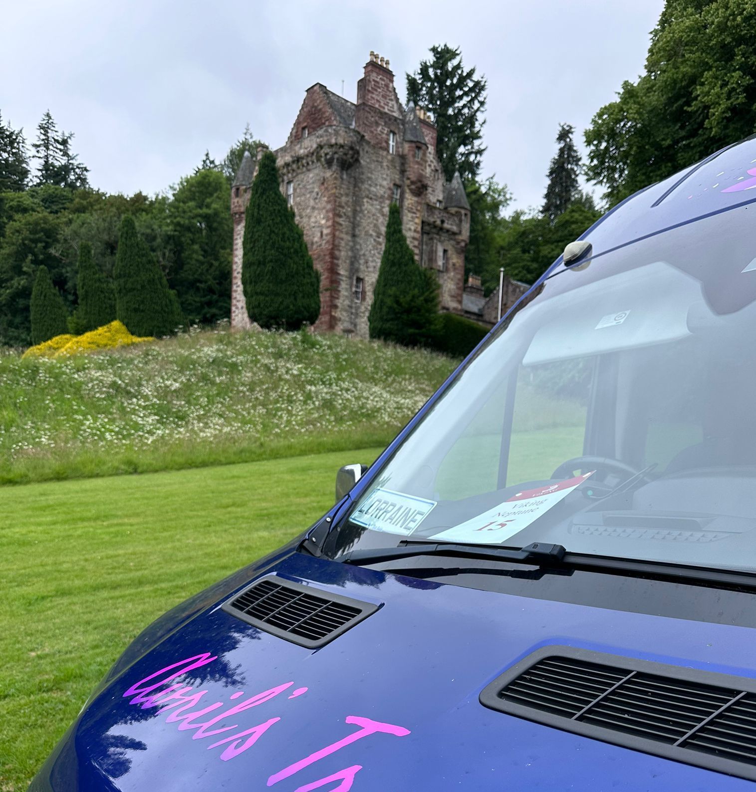 A blue van is parked in a grassy field with a castle in the background.