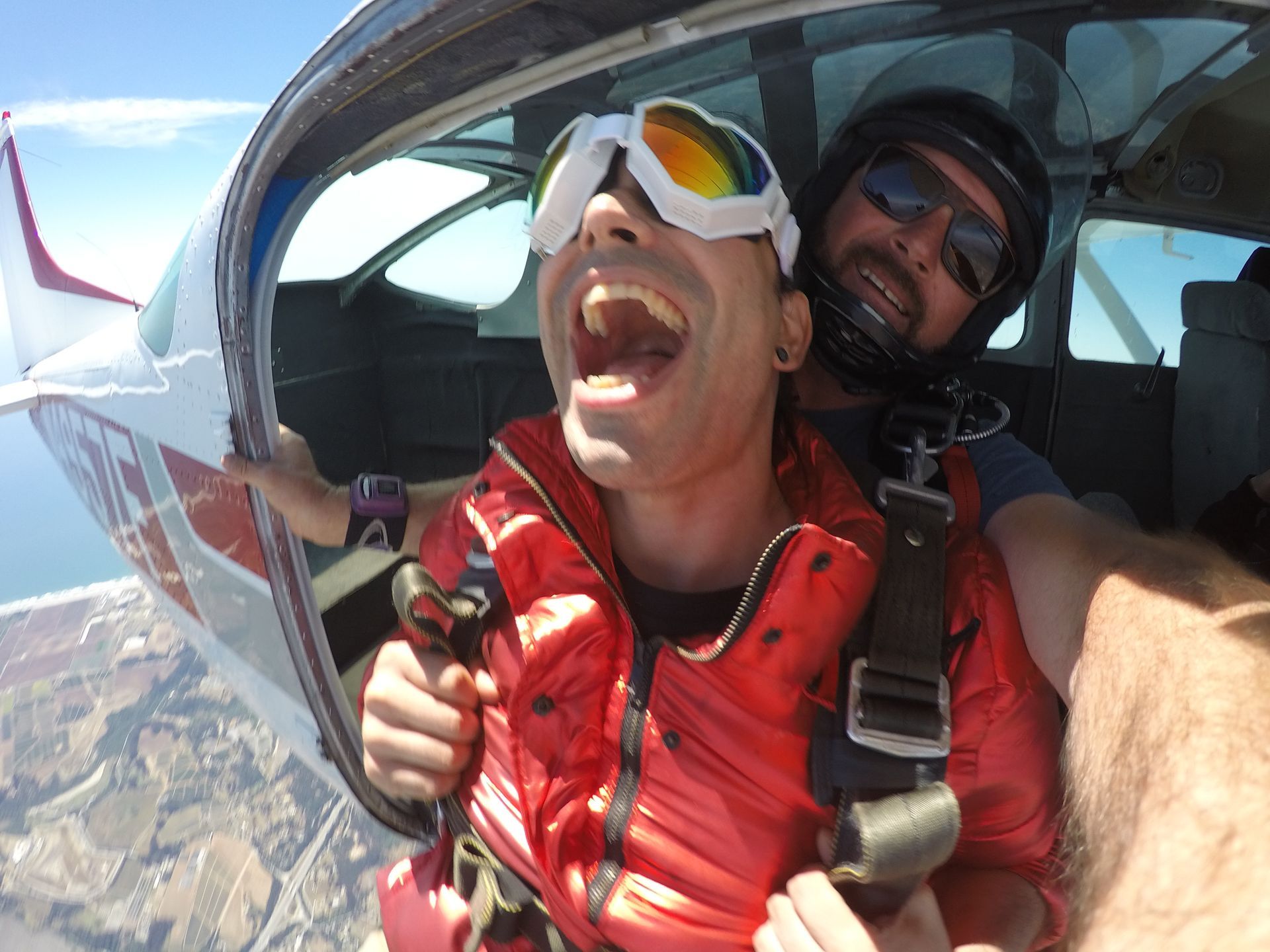 Tandem skydive pair prepare to jump from plane. Student has rainbow colored googles and has his mouth open with a big smile.