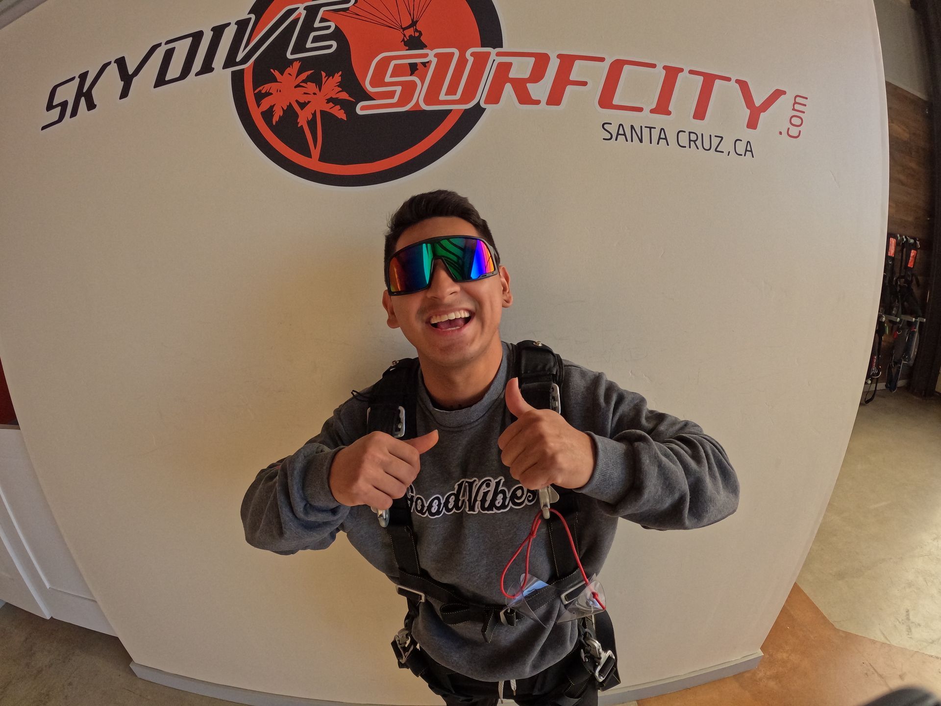 A man wearing sunglasses is giving a thumbs up in front of a sign that says Skydive Santa Cruz.