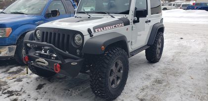 A white Jeep Wrangler Rubicon with a black aftermarket bumper and off-road tires parked on a snowy lot. | Hillman Tire Sales and Service LLC