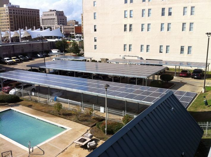 Solar panels on parking lot canopies over cars, next to a pool and building.