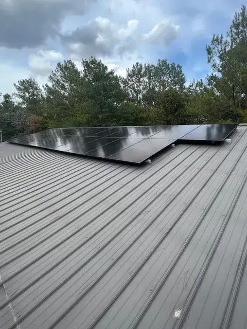 Solar panels installed on a gray corrugated metal roof with trees in the background under a cloudy sky.
