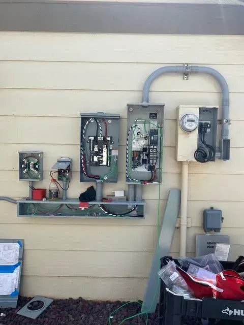 Electrical panel installation on a beige house exterior; various open boxes and conduit.