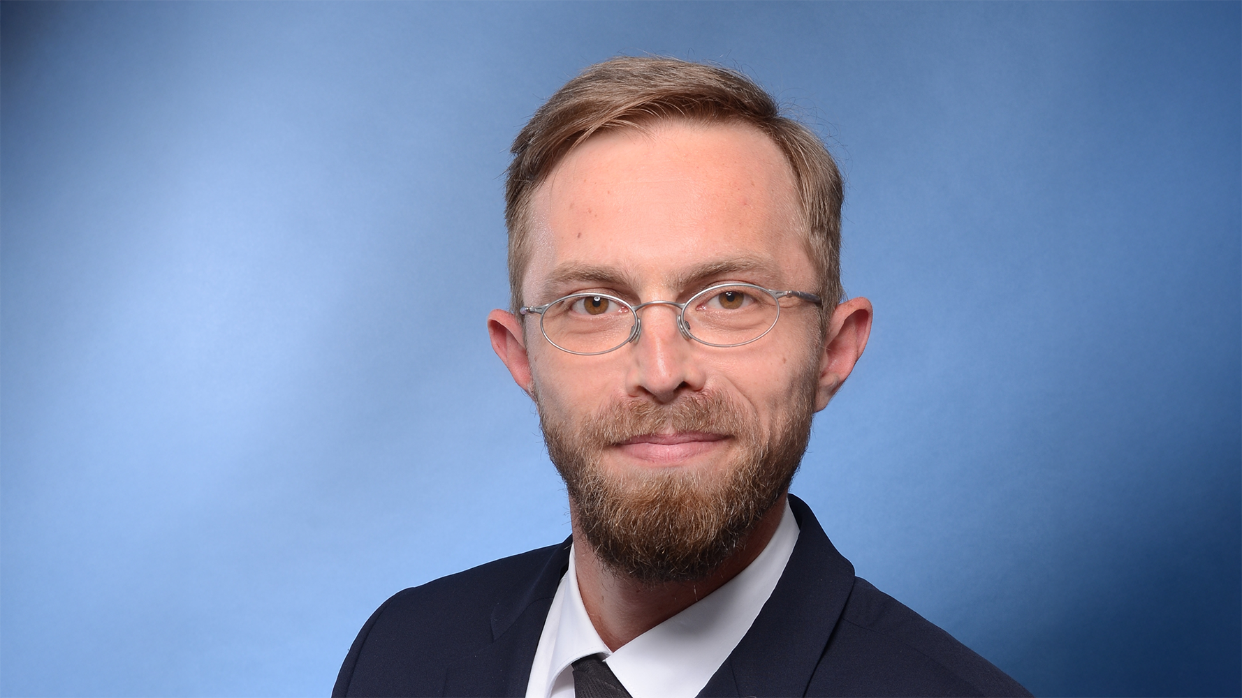 Mann mit Brille und Bart, der eine dunkle Jacke trägt, vor blauem Hintergrund.