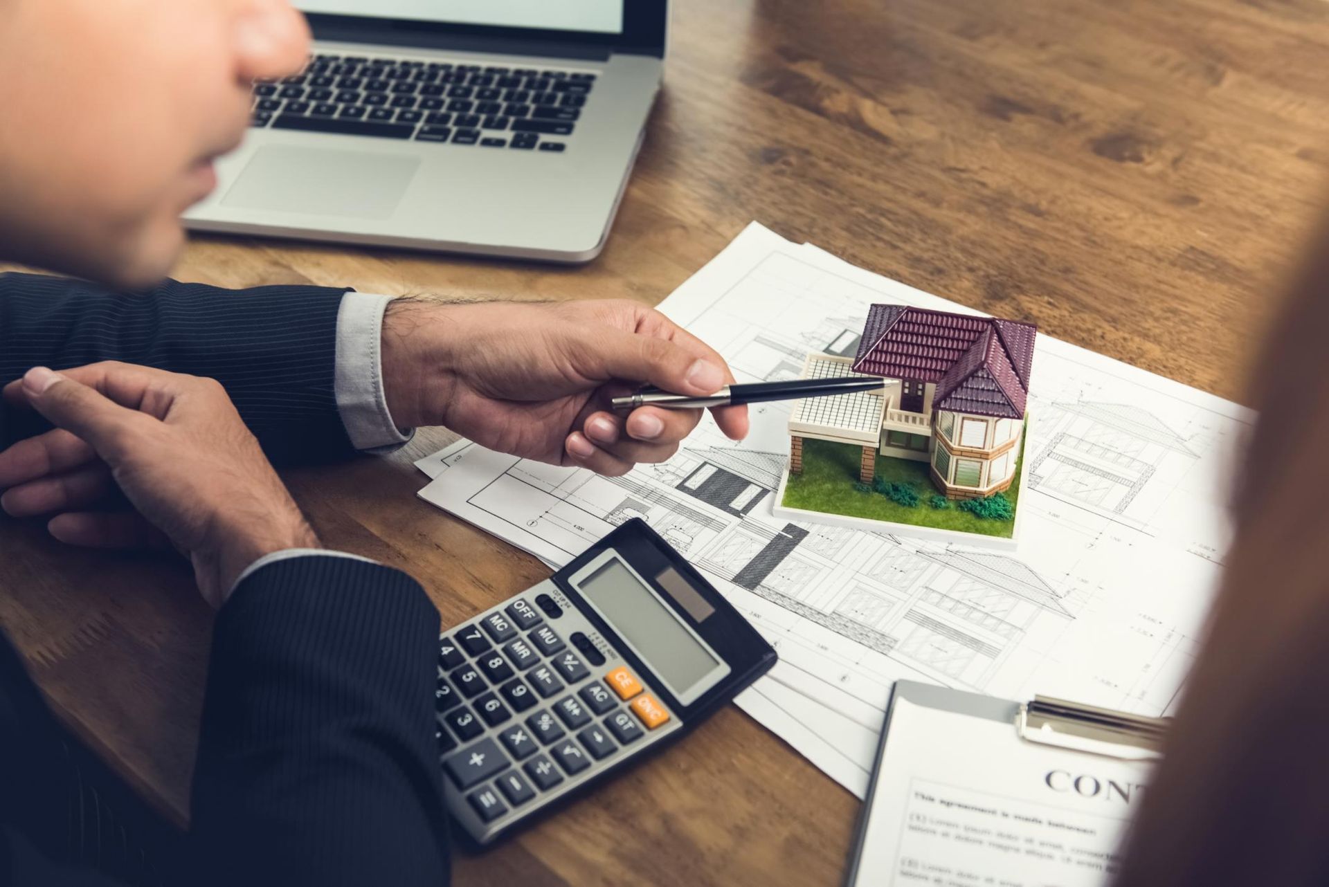 Person pointing at a miniature house on paperwork with a calculator and laptop, discussing real estate.