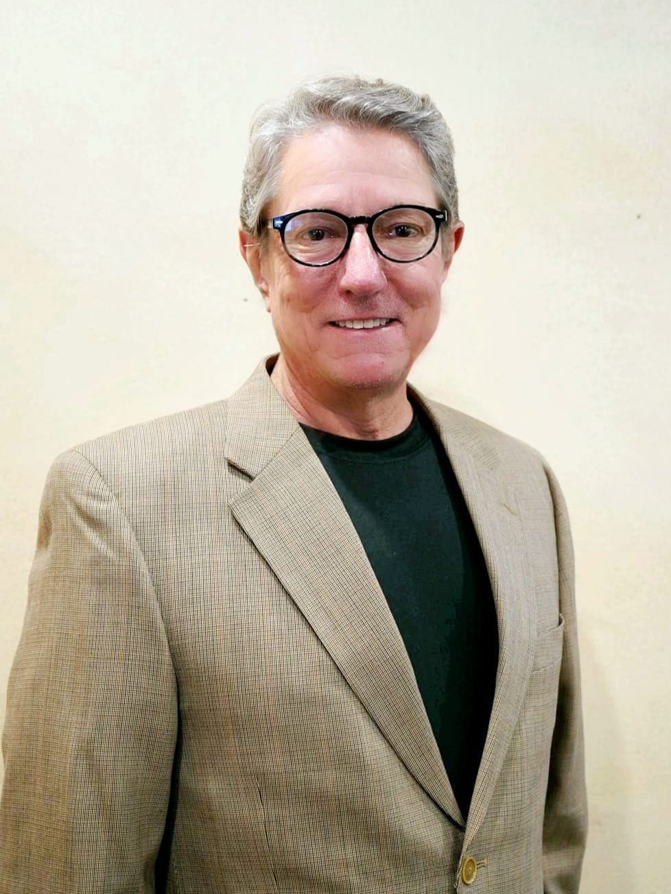 Matthew Everett in glasses, brown blazer over a dark top, smiling, neutral background.