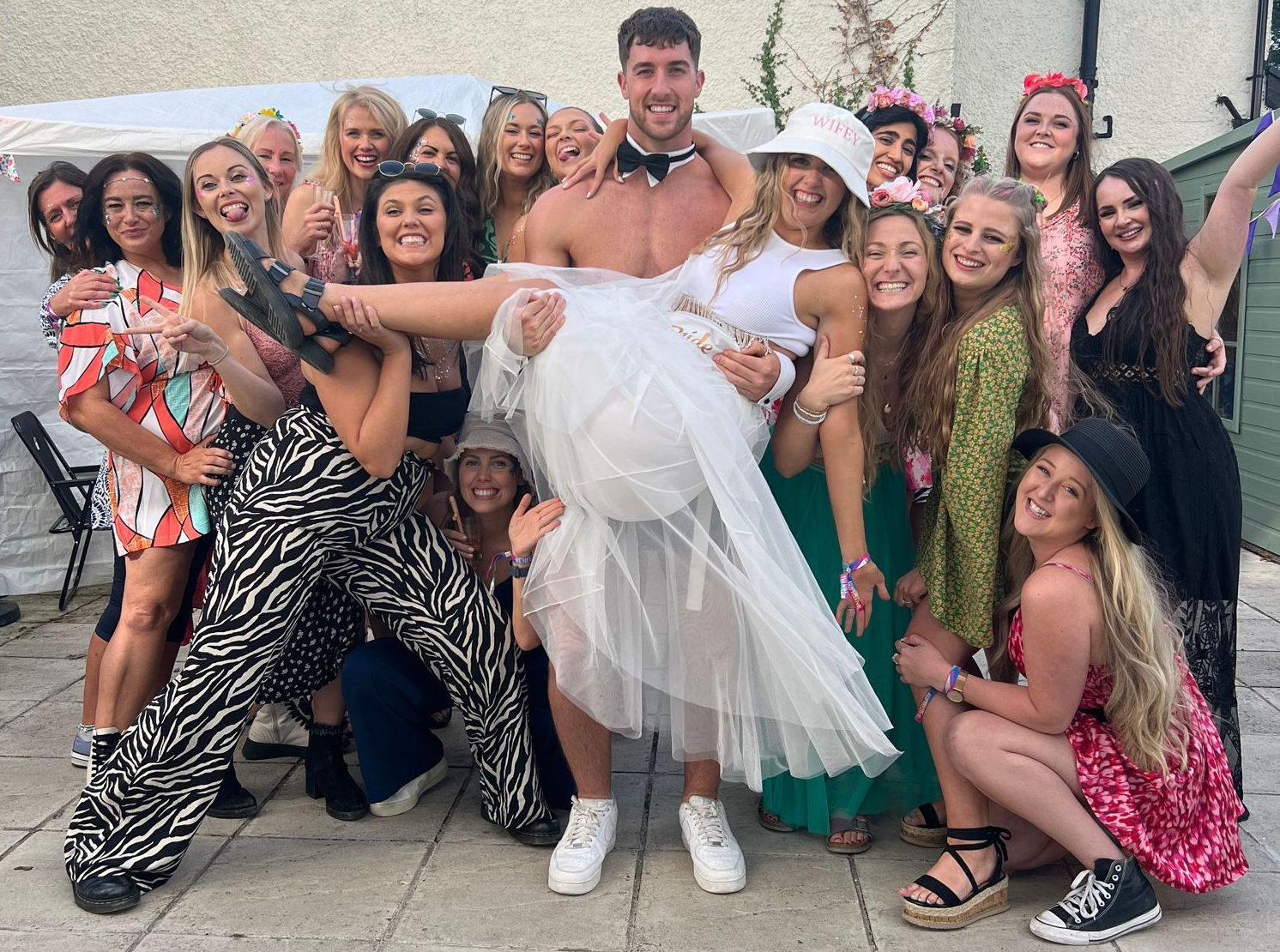Buff butler posing with a hen party group as a guest is held playfully during an outdoor celebration