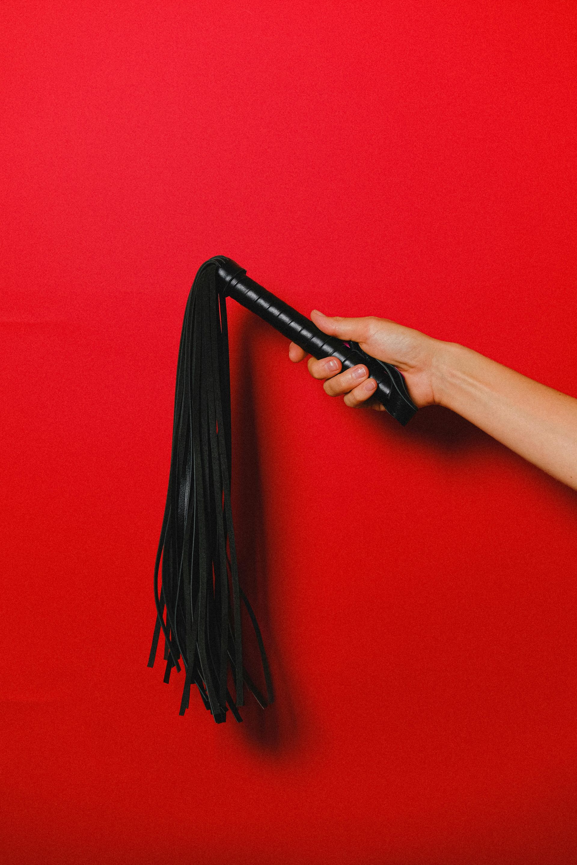 Close-up of a hand holding a black leather flogger against a bright red background, creating a striking contrast and a bold, dramatic visual tone