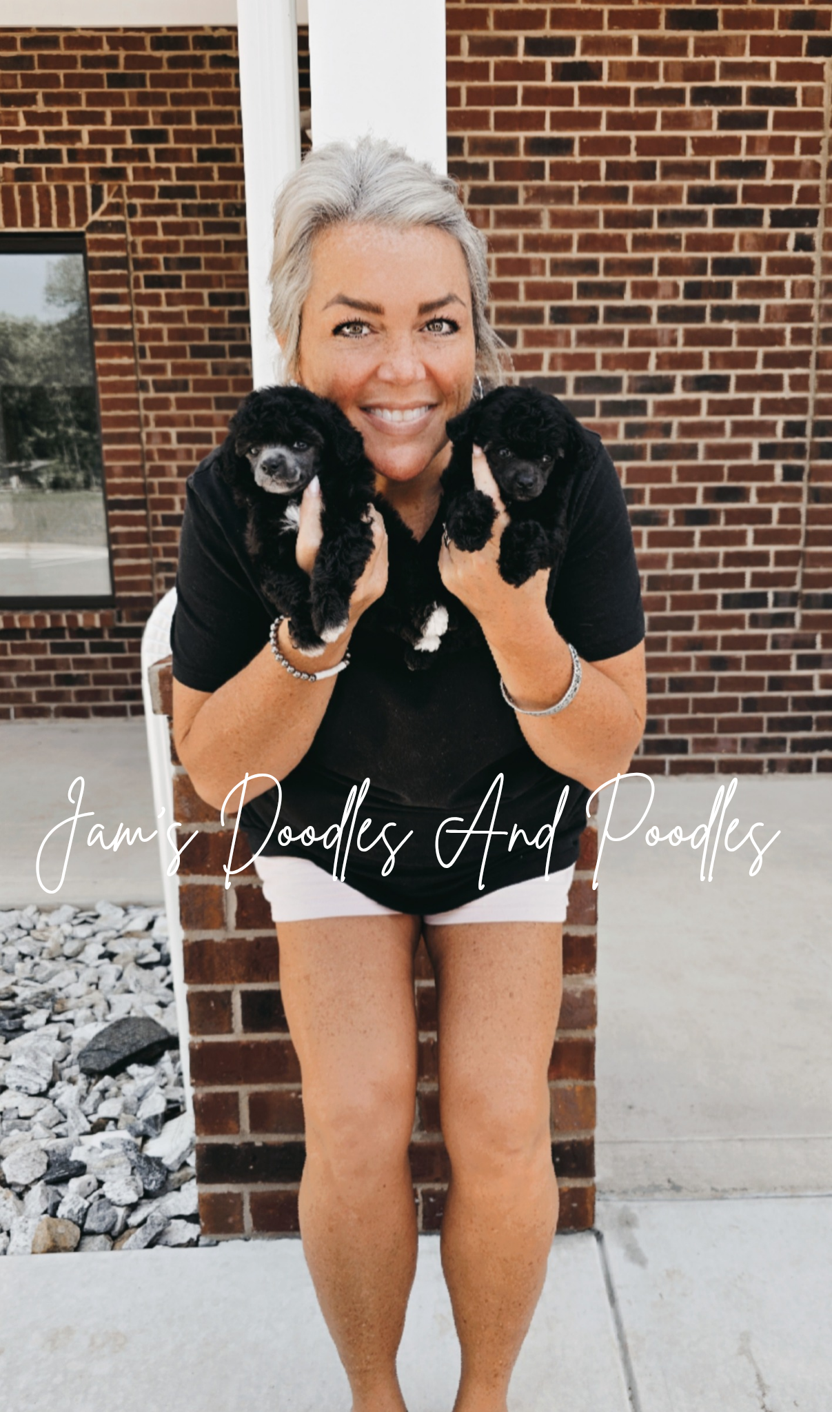 Woman smiling, holding two small black puppies. Brick building background.