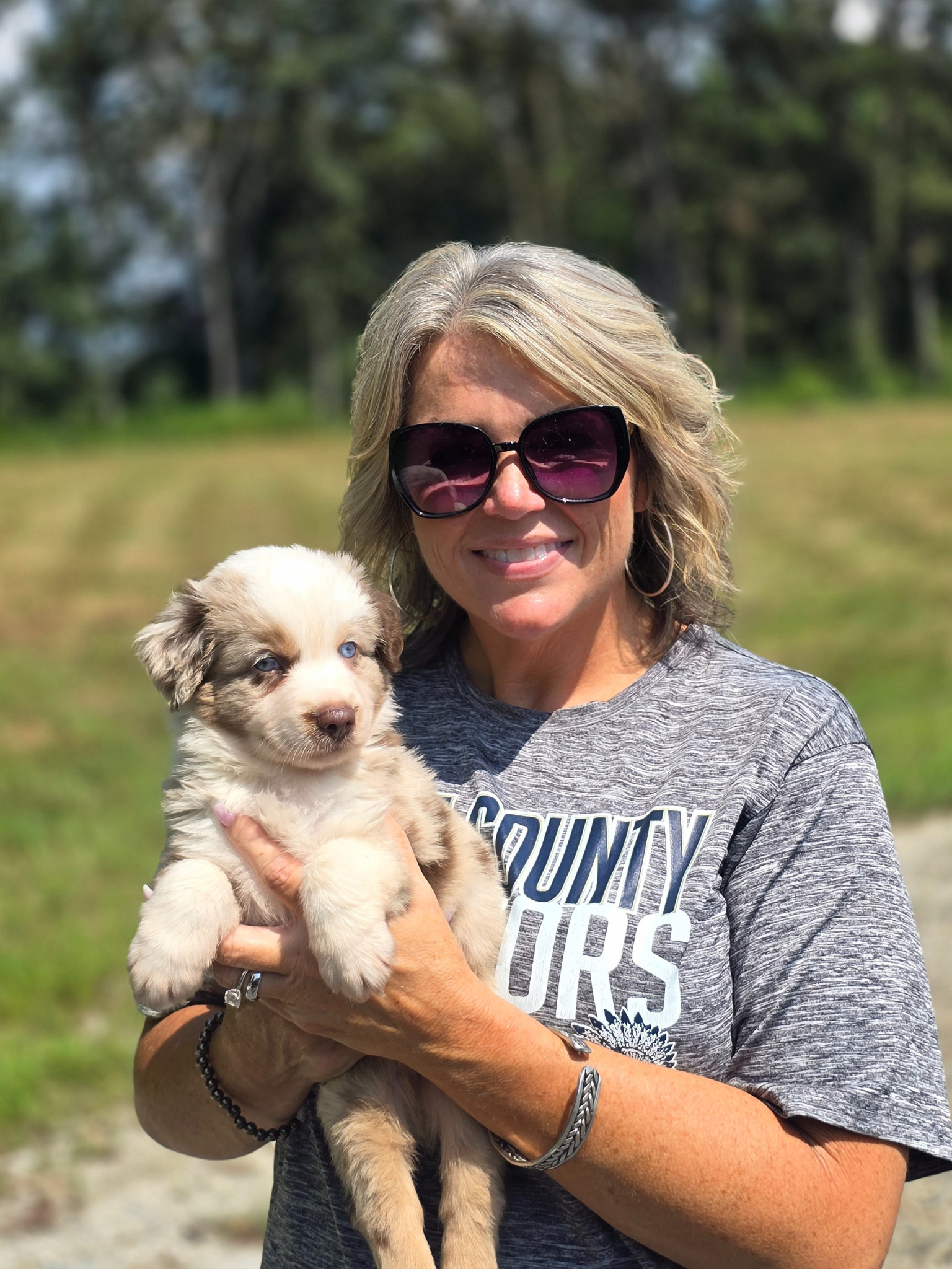 Woman holding a small, tri-color puppy outdoors, smiling, wearing sunglasses.