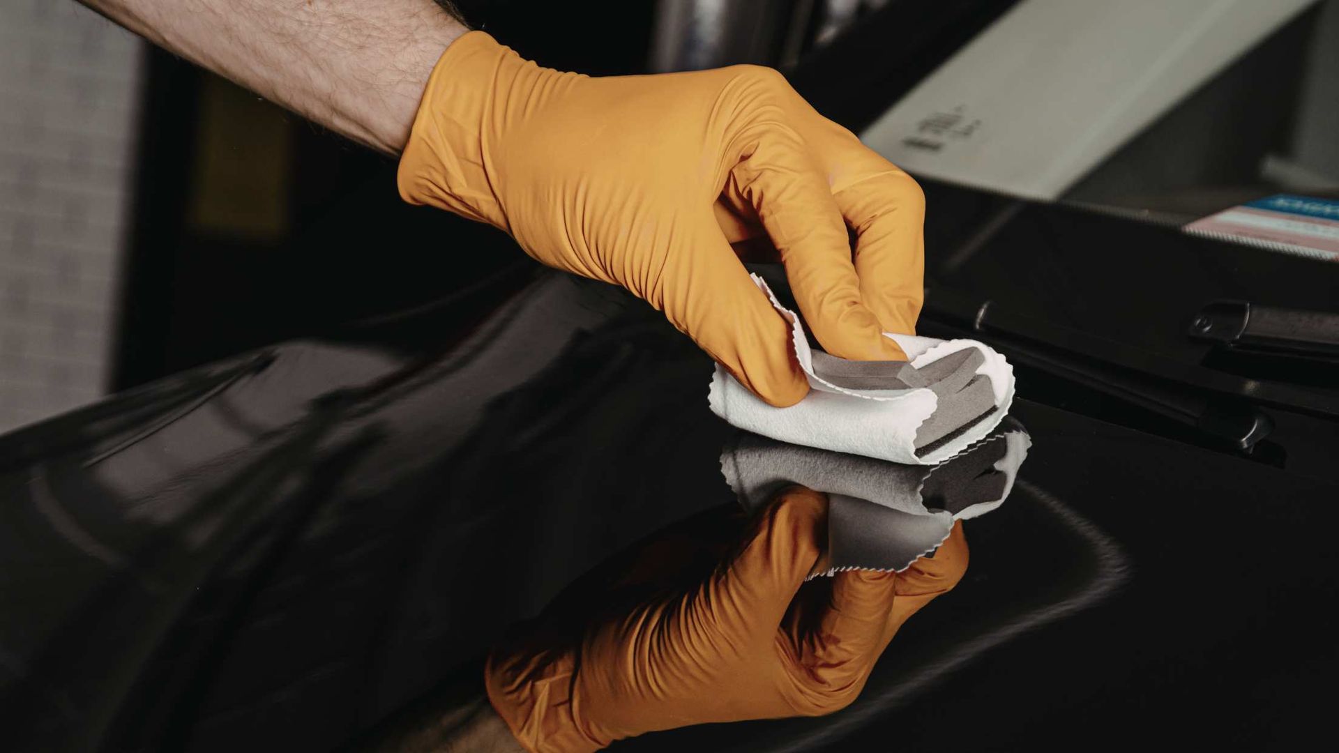 Gloved hand wiping a glossy black car surface with a cloth, reflecting the hand.