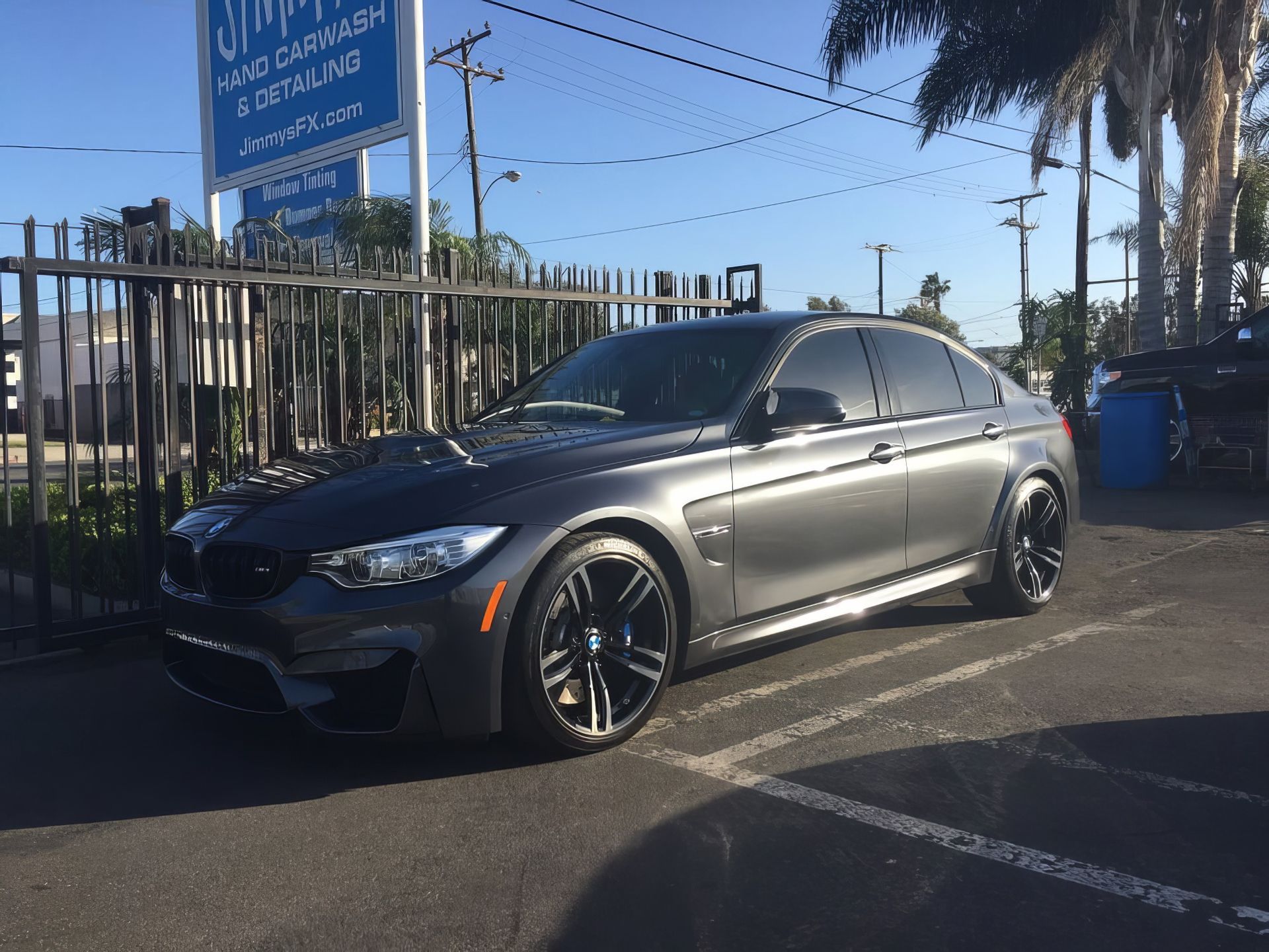 Silver BMW sedan parked on a sunny street beside a fence and palm trees