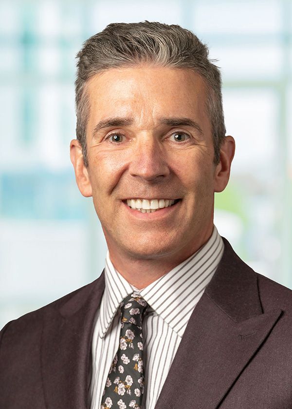 Portrait of a smiling male physician, David Miller, MD, wearing a suit jacket, striped dress shirt, and patterned tie, with a softly blurred clinical background, representing Retina Associates of Cleveland.