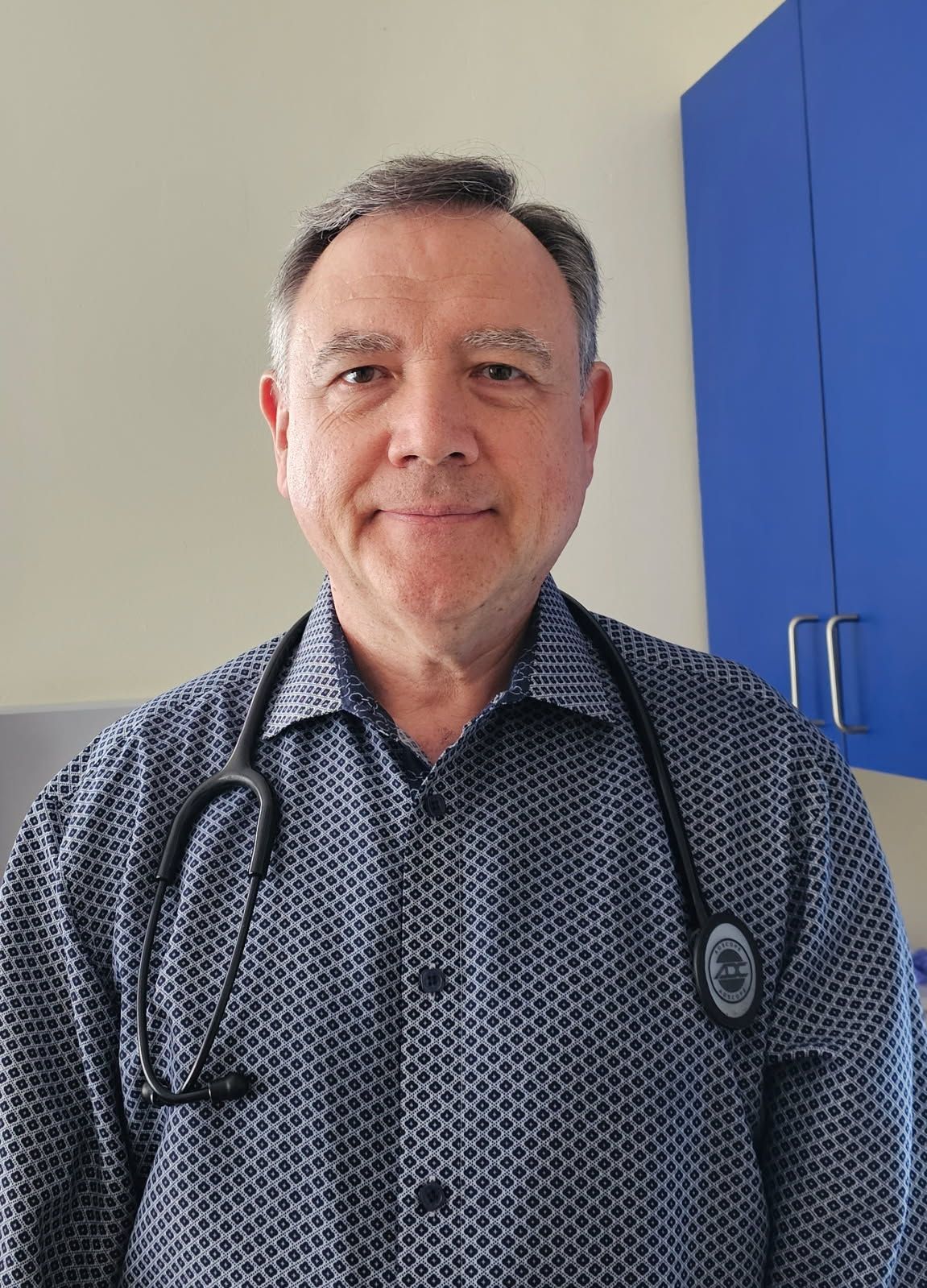 Dr. Martin Slavin, MD, a middle-aged man with short gray hair, is wearing a patterned navy shirt and a black stethoscope around his neck. He is standing indoors in a medical office, with a neutral-colored wall and blue cabinets in the background, looking directly at the camera with a slight smile.