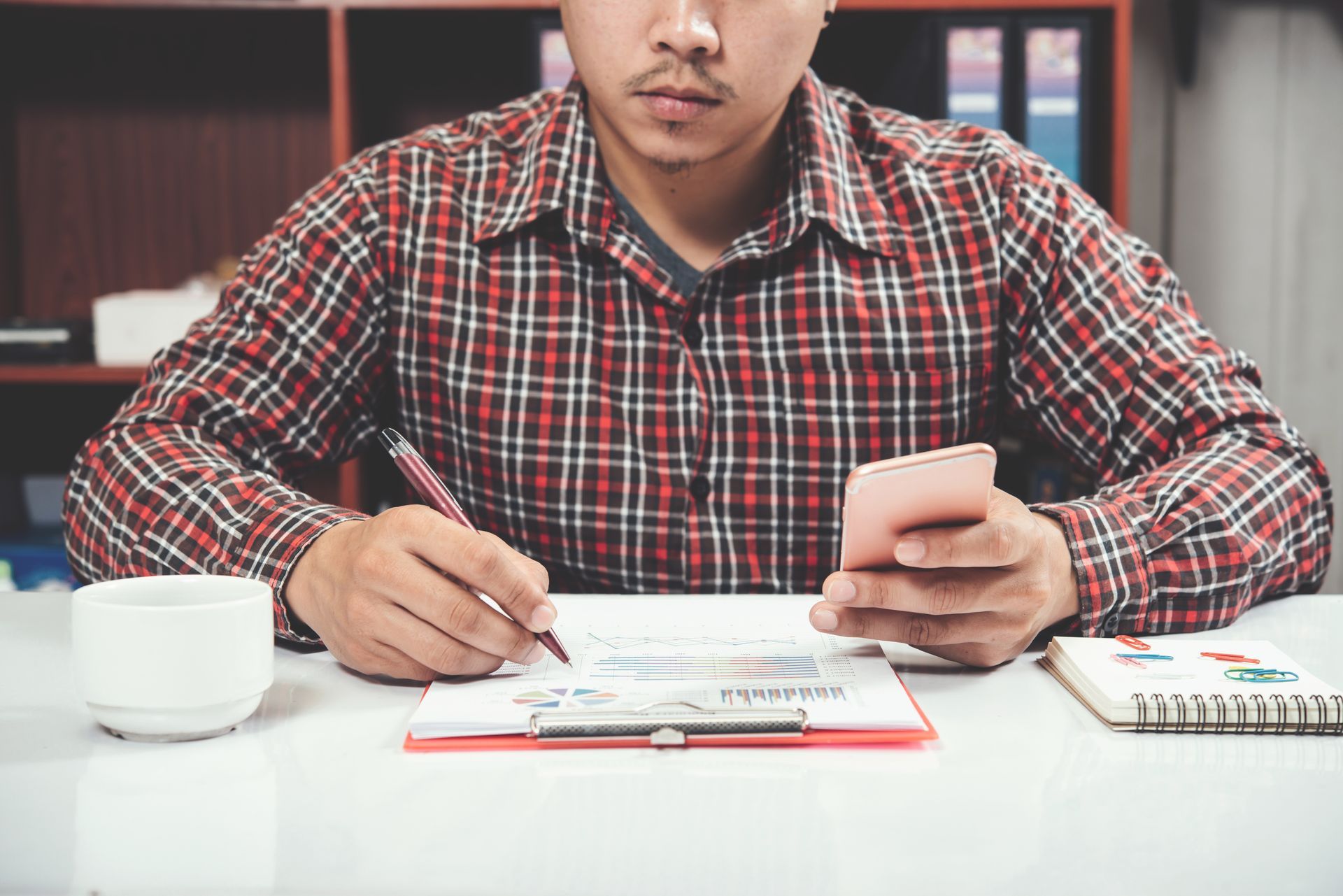 A man is sitting at a desk using a cell phone and writing on a clipboard.