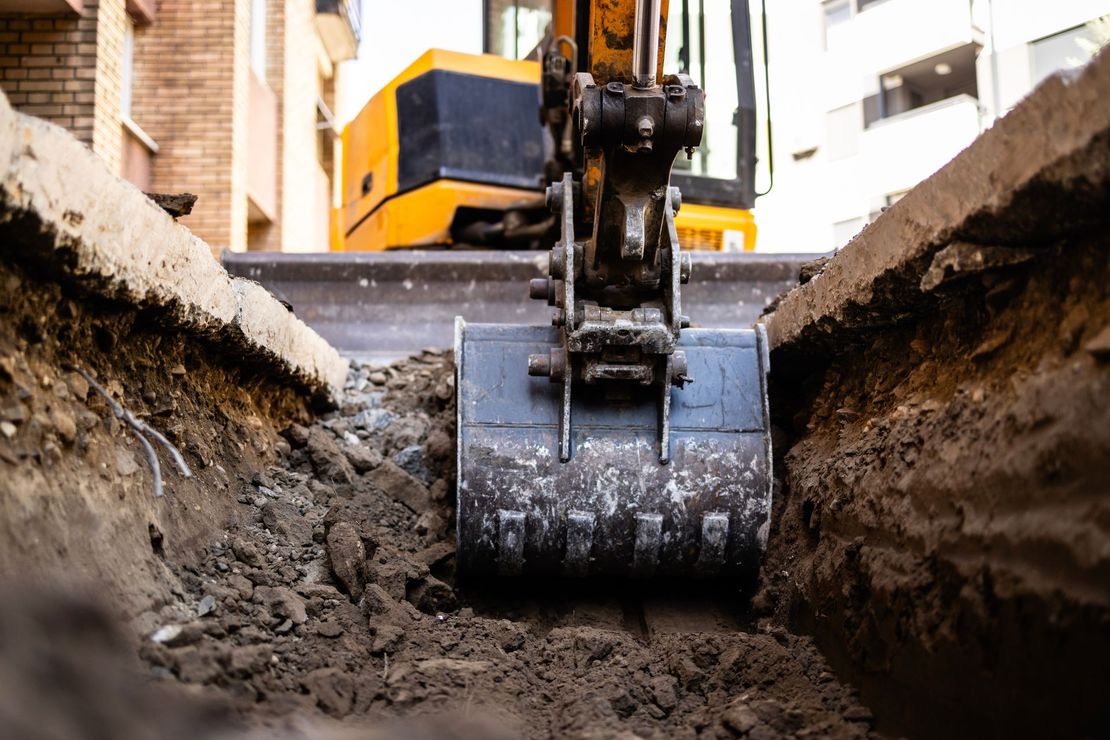 Yellow excavator bucket digging a trench. Dirt and concrete visible, with a building in background.