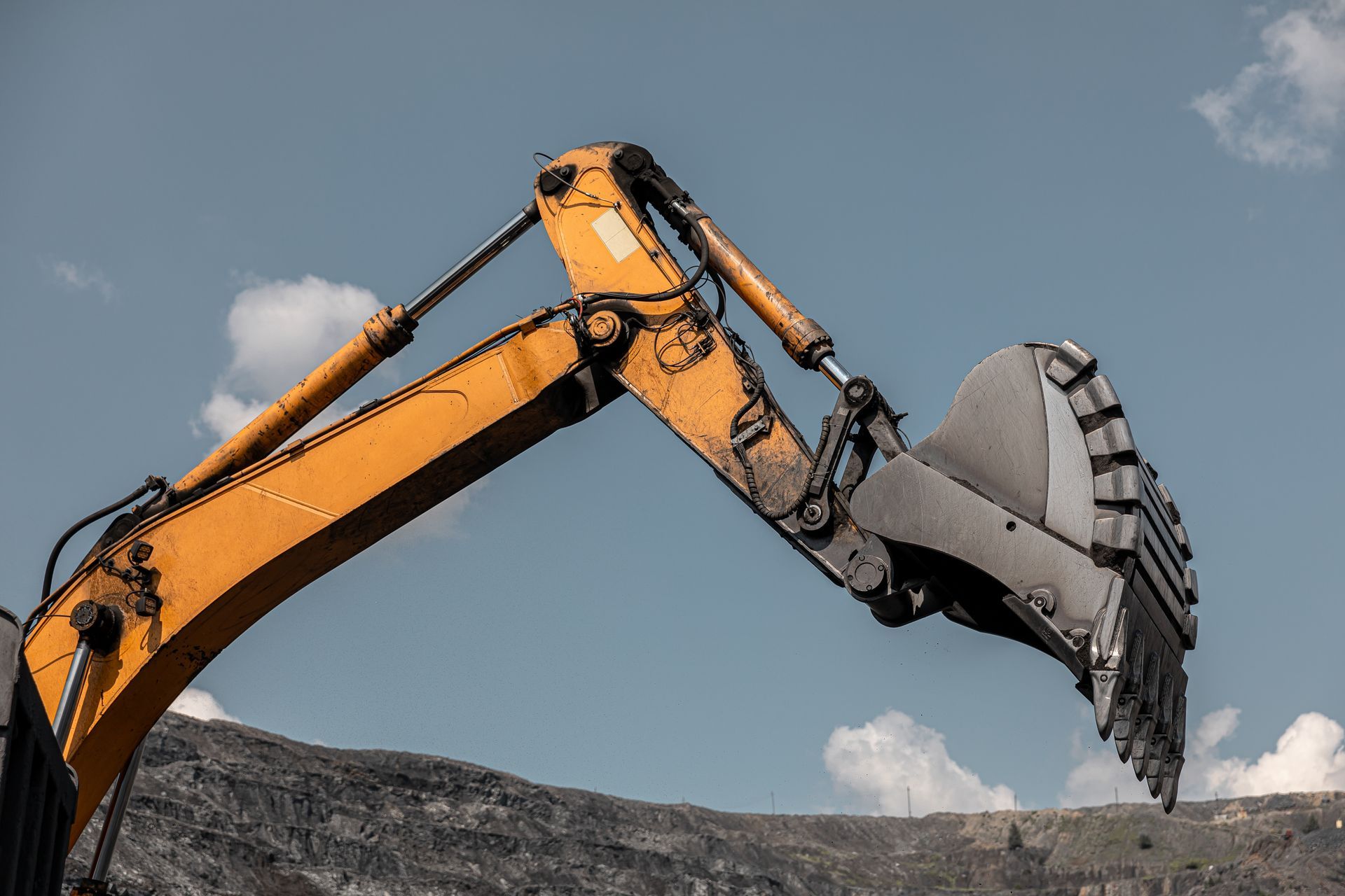 Yellow excavator arm and bucket against a cloudy sky, digging into a dirt pile.