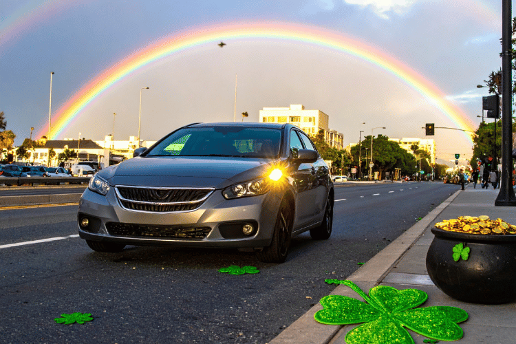 Sedan driving on a city street beneath a rainbow with shamrocks and a pot of gold nearby.