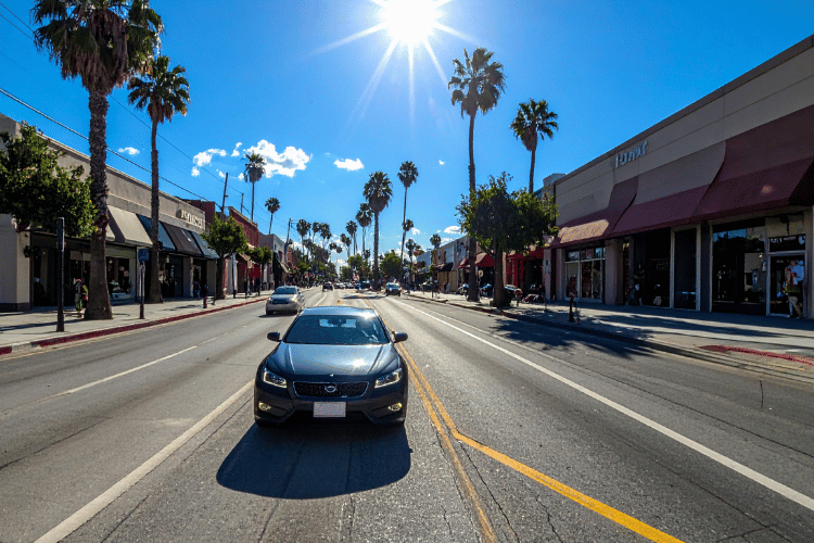 Black sedan driving down a sunny palm tree-lined street with storefronts and clear blue skies.