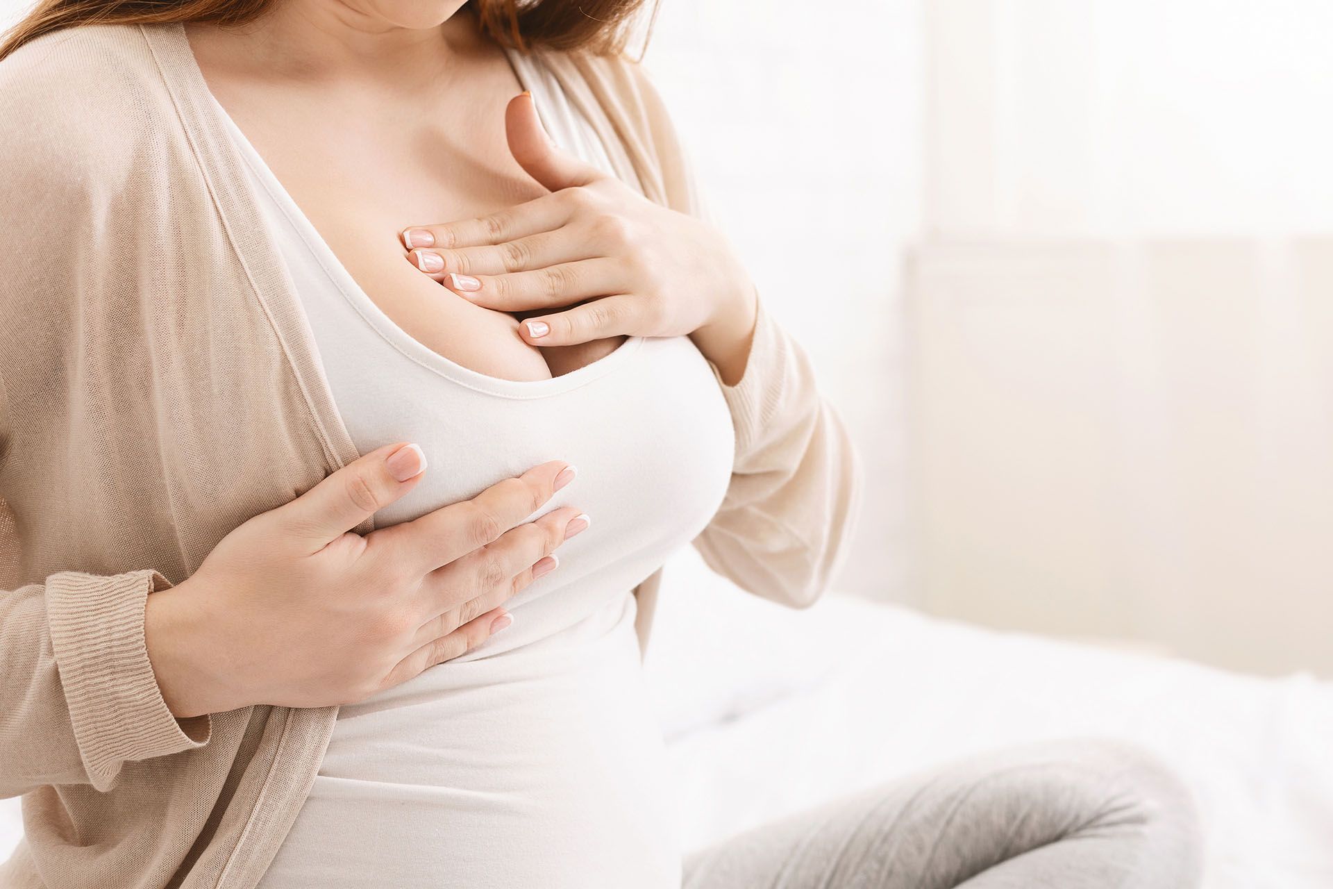 Woman touching her chest, possibly pregnant. Wearing white top and light cardigan. Sitting on a bed.