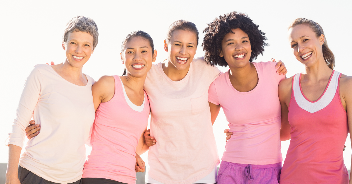 Five women in pink workout attire, smiling and embracing outdoors.