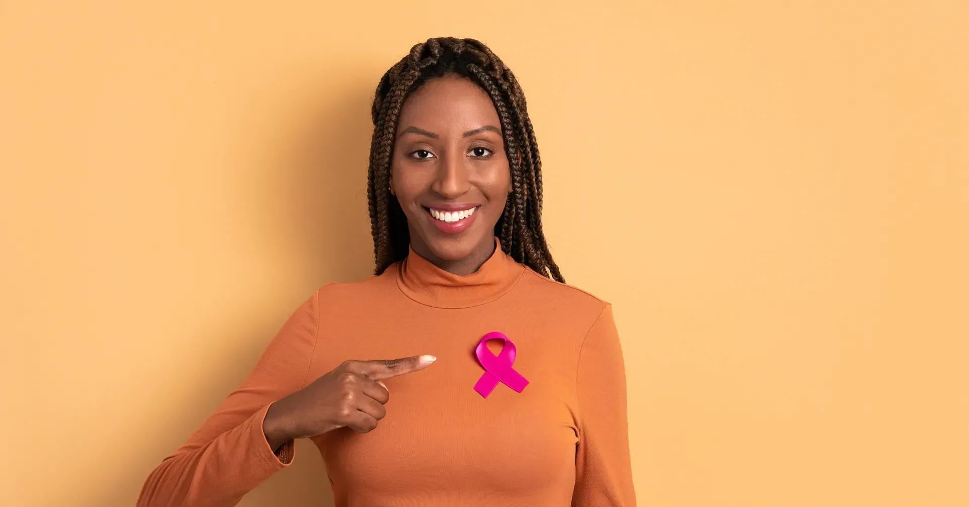 Woman with braided hair, smiling, pointing at a pink breast cancer awareness ribbon pinned to her shirt.