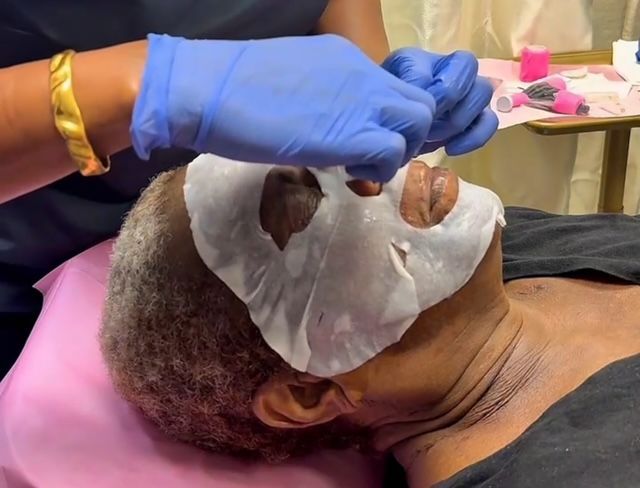 A person receiving a facial treatment. A technician in blue gloves applies a white sheet mask.