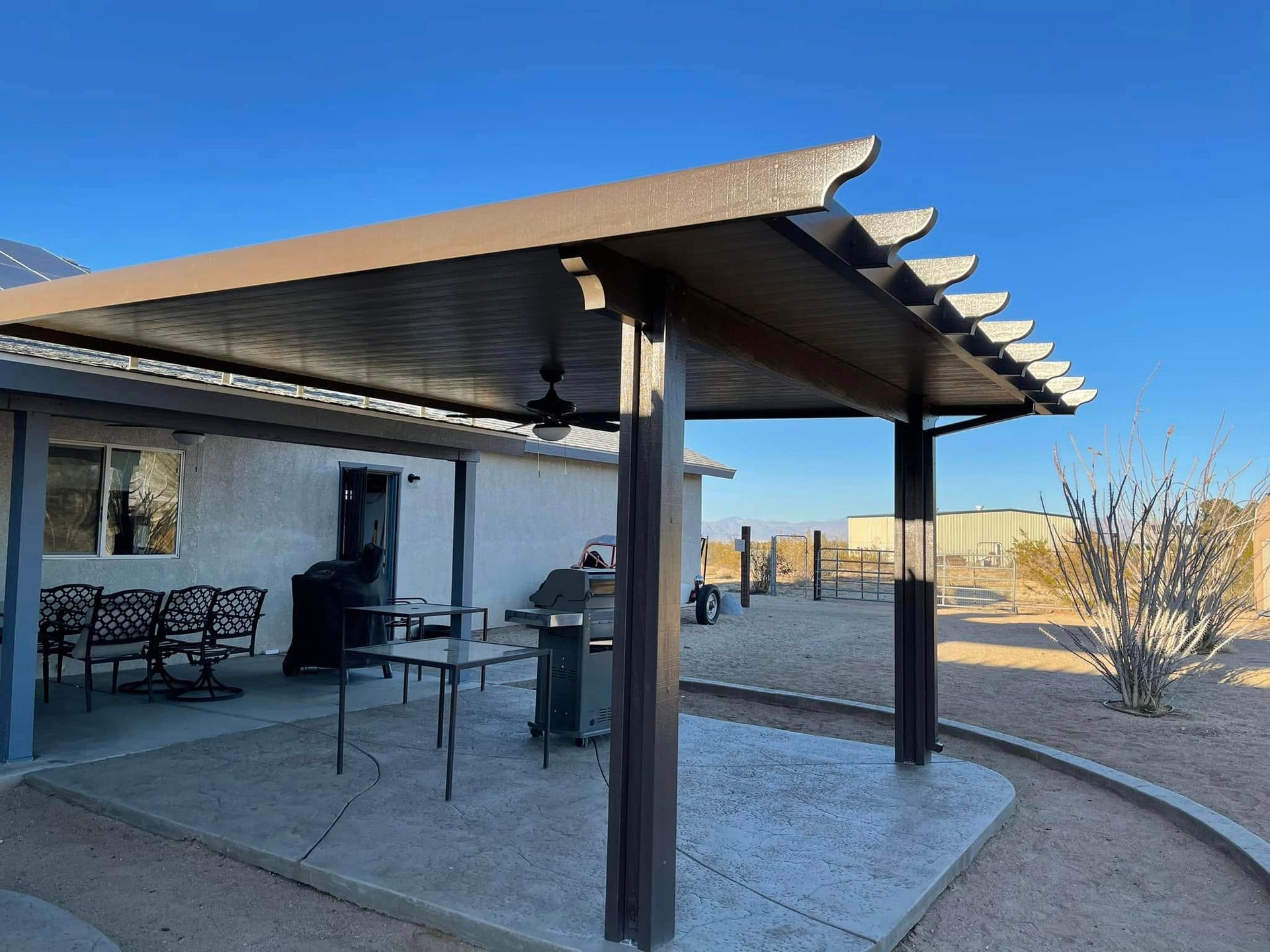 Patio cover attached to a home with grill, table, and chairs outside. Desert setting with blue sky.