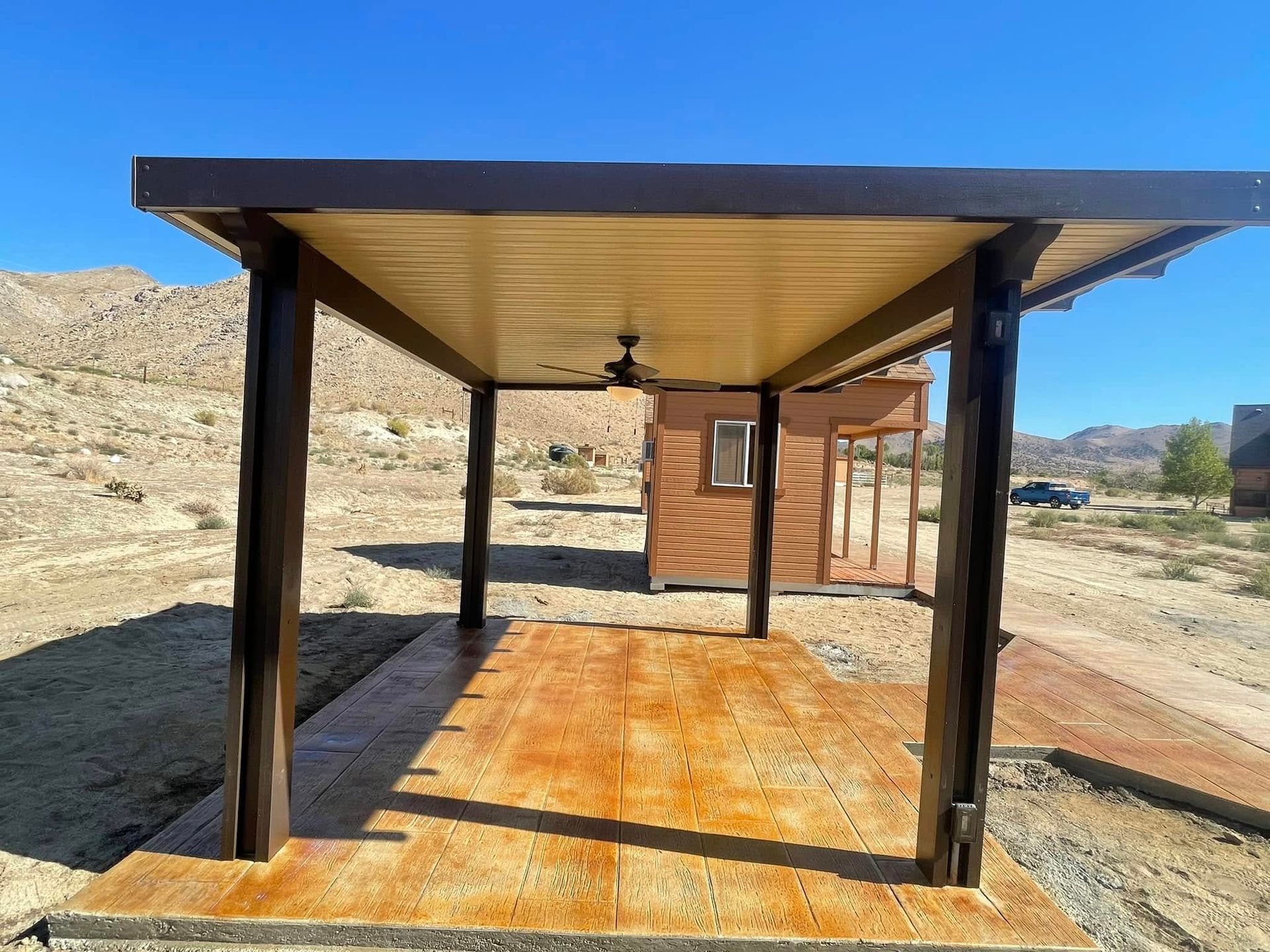 Covered patio with brown posts and ceiling, tan floor, and small wooden building in the background.