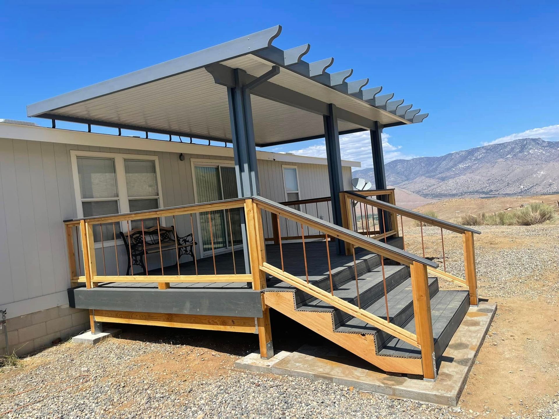 Deck and pergola attached to a light-colored building, steps leading up. Brown, grey, and blue colors. Mountains in the background.