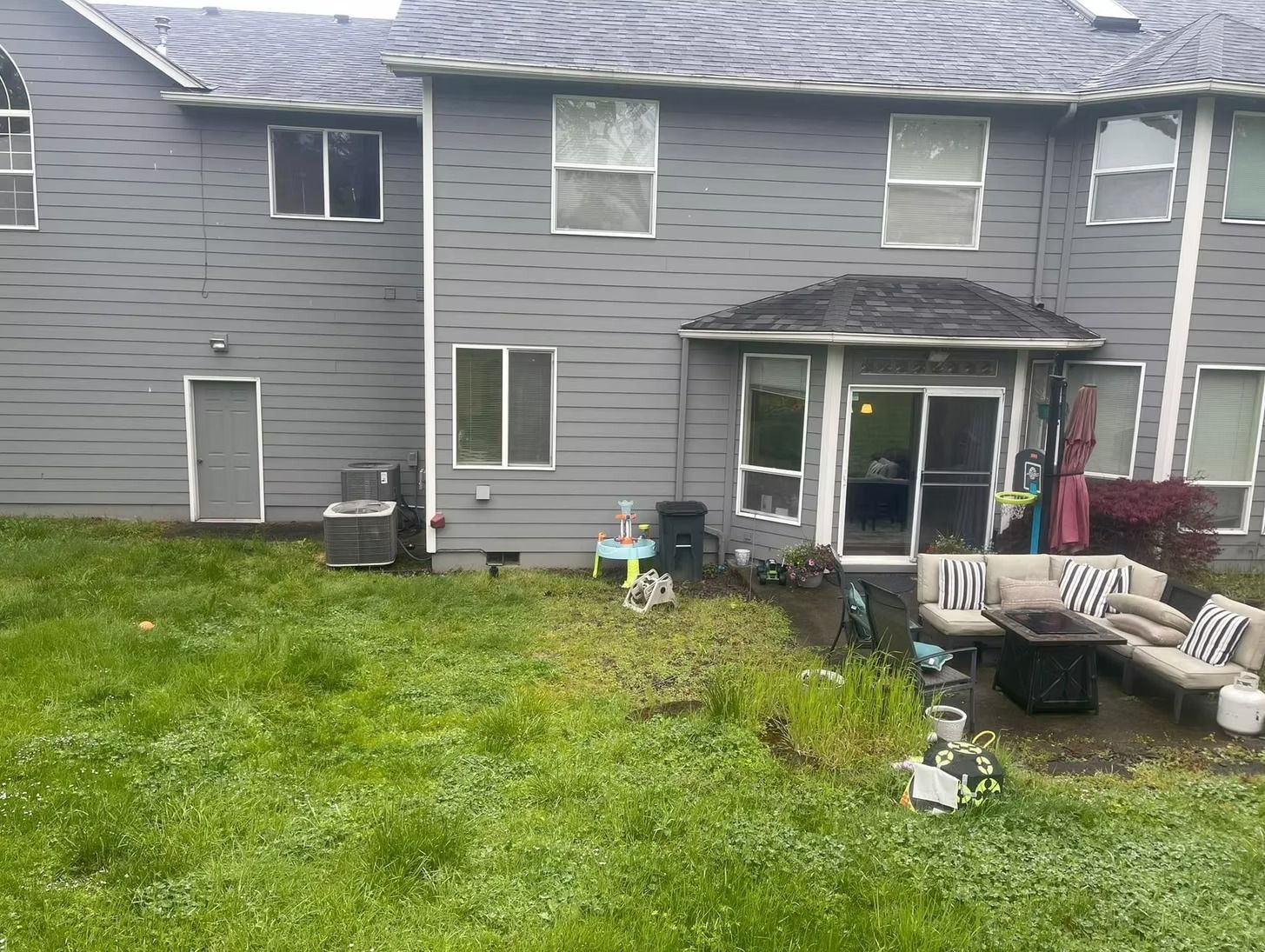 Backyard of a gray house with a patio and outdoor furniture, overgrown lawn.