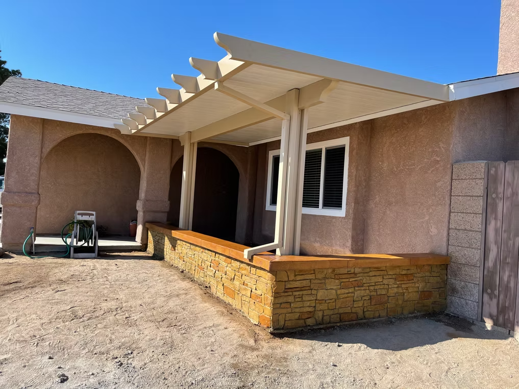 Tan stucco house with a pergola-covered porch, stone retaining wall, and bright blue sky.