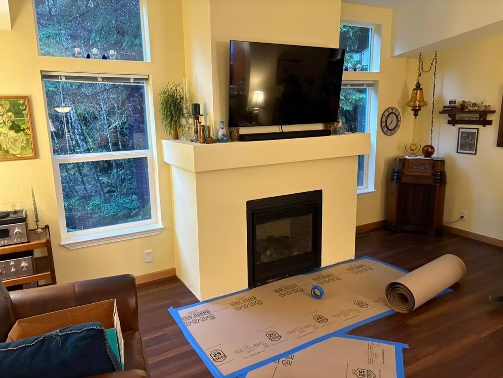 Living room with fireplace, TV above it. Brown paper protecting the floor.  Windows and dark wood flooring.
