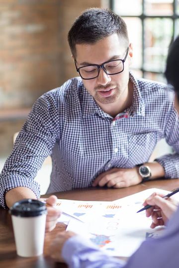 Man in glasses reviews paperwork with another person at a table, coffee nearby.
