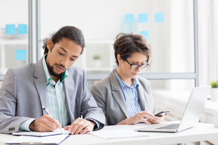 Two people working at a desk in an office. Man writing, woman using a phone and laptop.