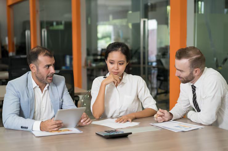 Three people in business attire at a table, discussing papers and a tablet in an office setting.