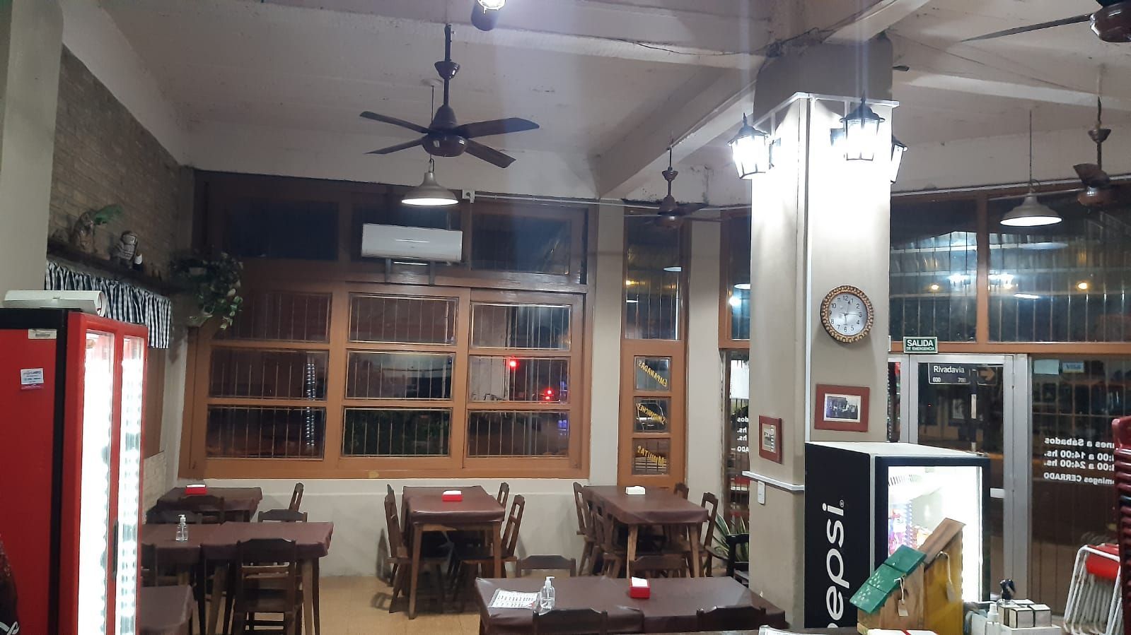 Interior de un restaurante con mesas y sillas de madera, ventiladores de techo y un refrigerador Pepsi.