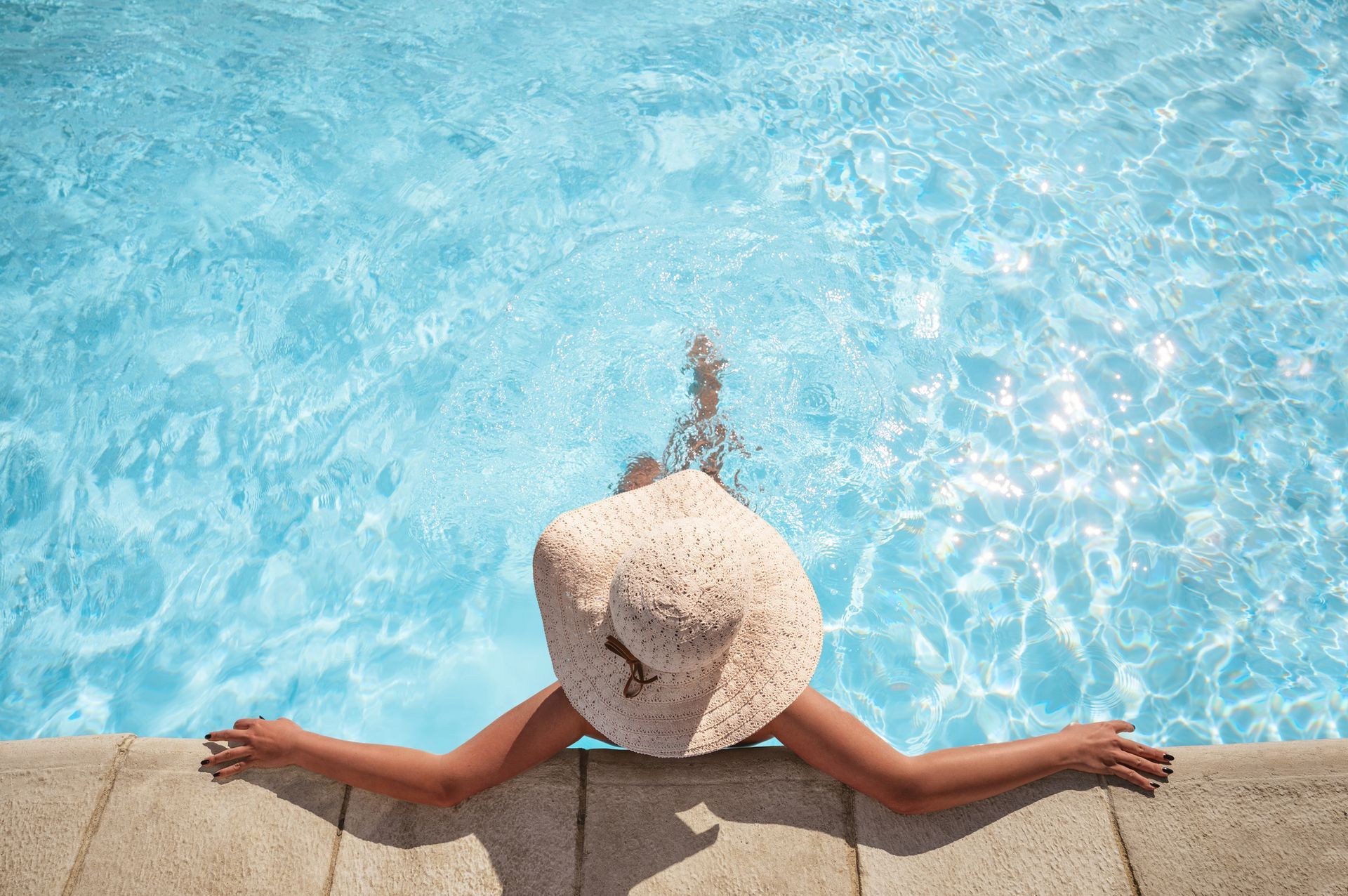 A woman in a hat is laying on the edge of a swimming pool.