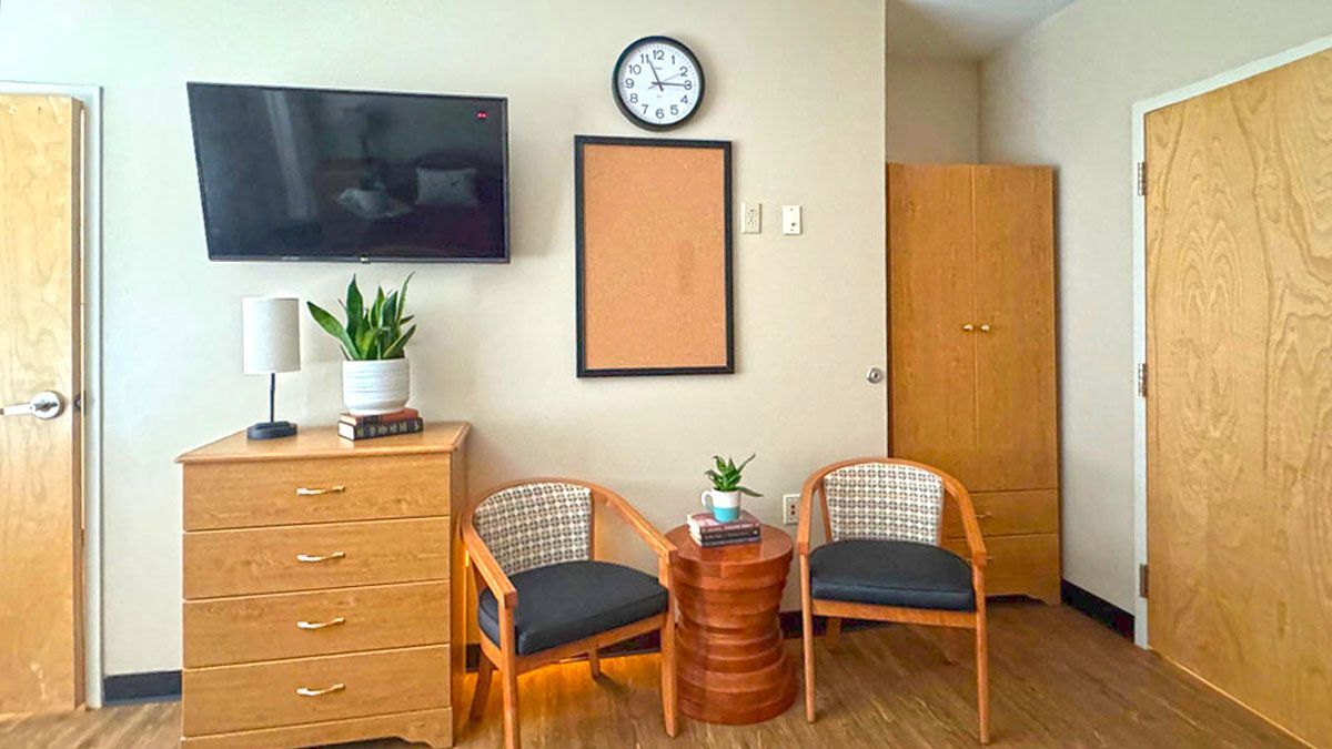 A living room with two chairs , a dresser , a flat screen tv and a clock on the wall.