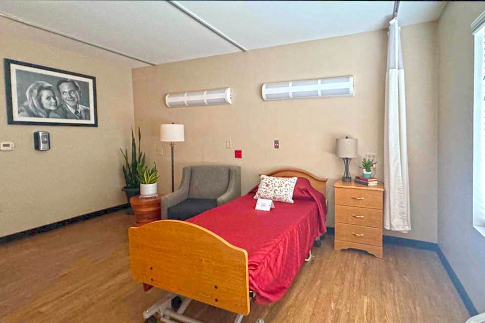 A hospital room with a bed , chair , nightstand and picture on the wall.