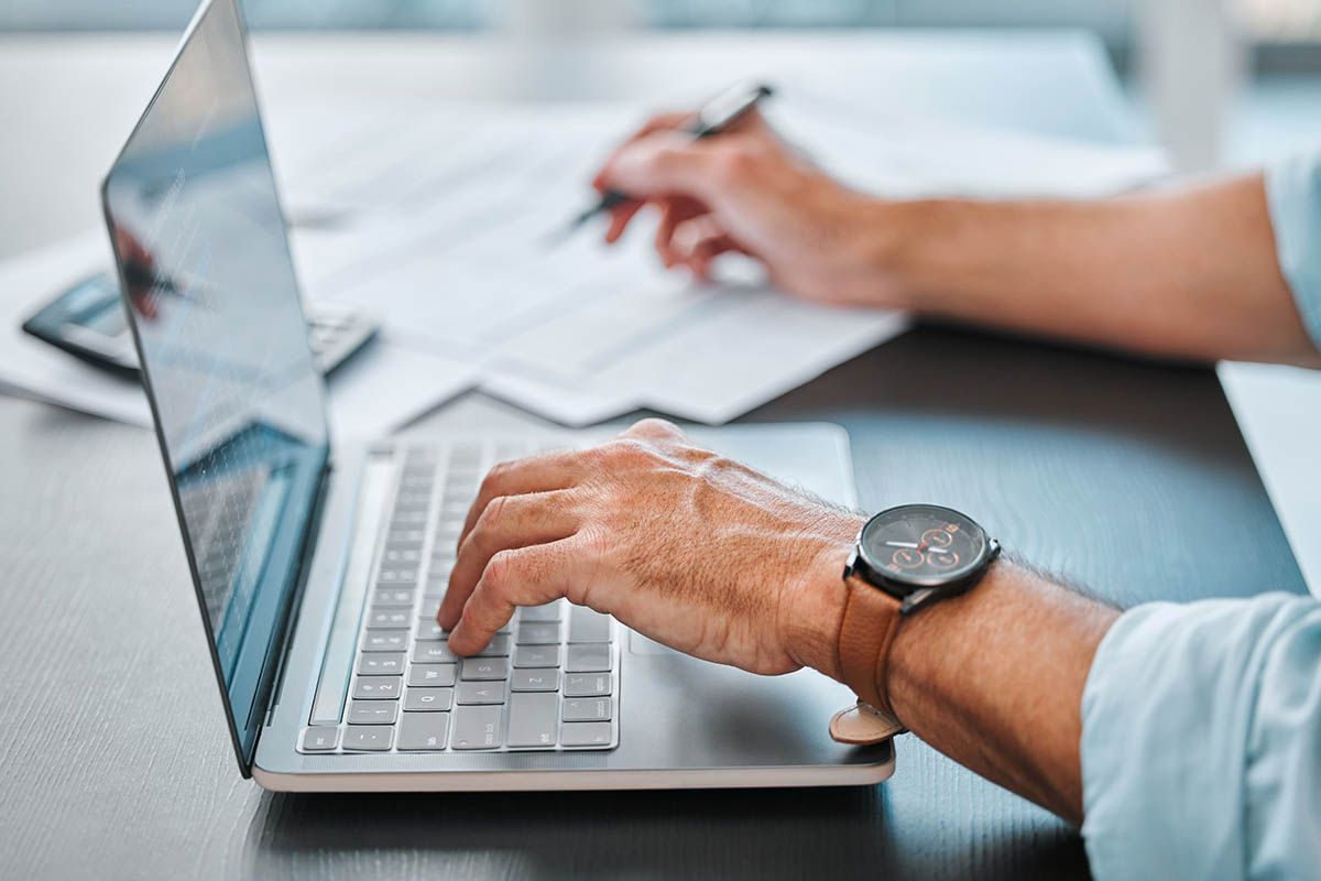 A man is typing on a laptop computer while writing on a piece of paper.